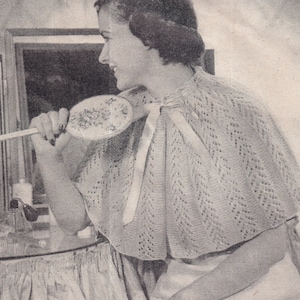 May include: A woman wearing a white crocheted shawl with a delicate lace pattern. The shawl has a white ribbon tied at the neck. The woman is holding a small, round, embroidered hand mirror.