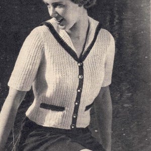 Può includere: Cardigan color crema a maniche corte con rifiniture scure intorno al colletto, alle tasche e all'abbottonatura. Il cardigan ha una trama a coste ed è abbottonato. I bottoni sono piccoli e rotondi. La persona indossa una gonna scura.