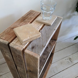 May include: A rustic wooden crate shelf featuring a weathered, light brown finish with visible wood grain. Two square, beige stone coasters are placed on the top surface, with a clear glass of water resting on one of the coasters.