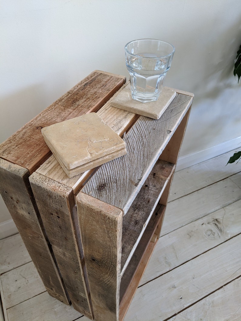 May include: A rustic wooden crate shelf featuring a weathered, light brown finish with visible wood grain. Two square, beige stone coasters are placed on the top surface, with a clear glass of water resting on one of the coasters.