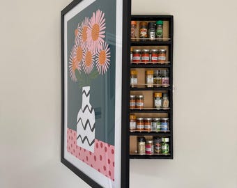 May include: A black-framed wall cabinet featuring a hinged door with a print of pink flowers in a white vase with black zig-zag patterns on a pink polka-dot surface. The interior contains six shelves holding various small glass spice jars.