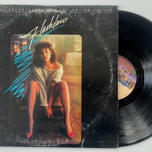 May include: A vintage vinyl record album for the movie "Flashdance." The album cover features a woman in a gray sweater and red heels, with the title in a stylized font and a turquoise design. The record is black.