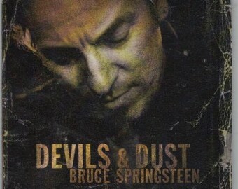 May include: Square drinks coaster with a close-up of a man's face in yellow and green tones. The text "DEVILS & DUST" and "BRUCE SPRINGSTEEN" are printed in gold on a dark background, likely a music album cover.