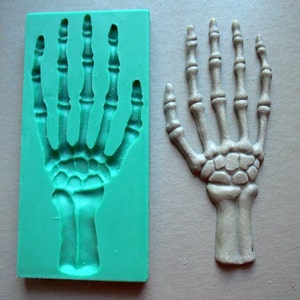 May include: A teal silicone mold and a beige skeleton hand. The mold is rectangular with a hand-shaped cavity. The hand is detailed with visible bones and joints, perfect for Halloween or crafting projects.