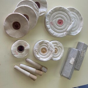 May include: An assortment of buffing wheels and polishing compounds. The buffing wheels are white, circular, and have a central brown or pink core. Several cylindrical buffing sticks and two wrapped rectangular bars are also present.