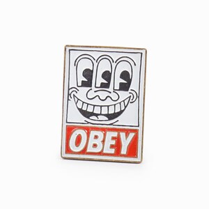 Könnte beinhalten: Eine weiße Emaille-Anstecknadel mit einem schwarz-weißen Cartoon-Gesicht mit drei Augen und einem breiten Lächeln. Das Gesicht befindet sich in einem weißen Quadrat mit schwarzem Umriss. Das Wort "OBEY" ist in roten Buchstaben auf weißem Hintergrund unter dem Quadrat gedruckt.