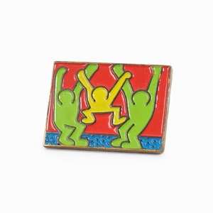 Pode incluir: Um broche de esmalte colorido com três figuras de pau em uma pose brincalhona. As figuras são verdes, amarelas e vermelhas sobre um fundo vermelho com detalhes azuis e brancos.