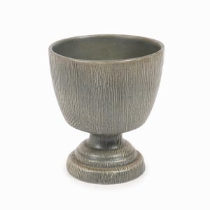 Puede incluir: Un cuenco gris de cerámica sobre pedestal con una apariencia texturizada similar a la madera. El cuenco tiene una parte superior ancha y abierta y se asienta sobre una base corta y redonda. El cuenco mide unos 15 cm de alto.