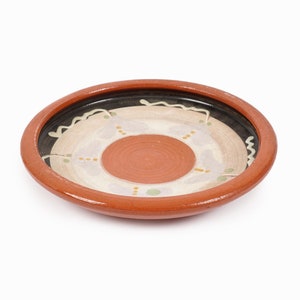 May include: A terracotta plate with a white and brown painted design. The plate has a raised rim and a circular design in the center.