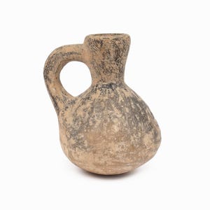 Puede incluir: Jarra pequeña de cerámica de estilo antiguo, con cuerpo redondeado y asa. La jarra es principalmente de color beige con detalles en gris oscuro y negro, lo que le da un aspecto envejecido. El asa tiene una abertura circular.