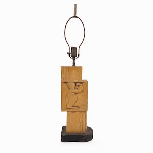 May include: A tan-colored table lamp with a carved relief of a person. The lamp has a rectangular base and a stacked design. The lamp's metal hardware includes a bulb socket and a lamp shade holder.