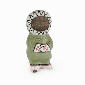 May include: A ceramic figurine of a person wearing a green hooded coat with red and white accents. The person has a brown face and a white hood with black markings.