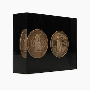 Könnte beinhalten: Eine schwarze rechteckige Plakette mit zwei bronzenen Medaillons. Das linke Medaillon zeigt den Text "The Mutual Life Assurance Society of the United States 1859" und eine Darstellung von zwei Figuren. Das rechte Medaillon zeigt den Text "1974 115th Anniversary" und eine Darstellung einer einzelnen Figur.