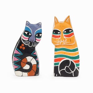 May include: Two hand-painted wooden cat figurines. One is black with blue, pink, and orange accents, and the other is orange with green and red stripes. Both have stylized features and a whimsical design.