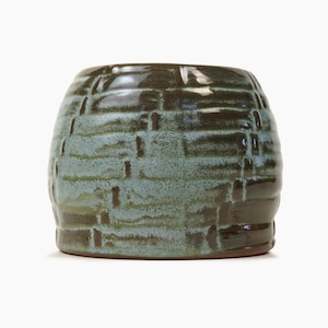 Op de afbeelding: Een ronde, groene keramische pot met een geweven patroon. De pot heeft een lichtgroene glazuur met bruine accenten.