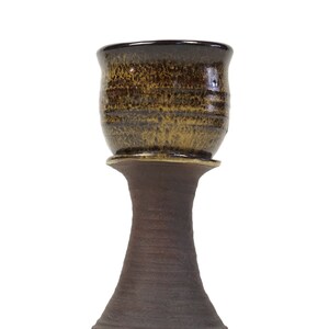 May include: A brown ceramic cup with a flared rim sits on a matching brown pedestal. The cup has a speckled glaze with a darker brown rim.
