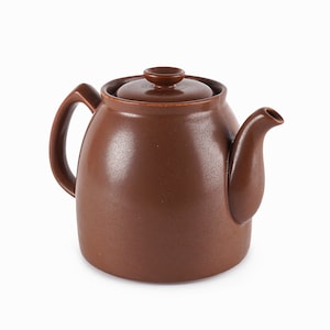 Puede incluir: Una tetera de cerámica marrón con tapa y asa curva. La tetera tiene un pico y un cuerpo redondeado. La tetera es de color marrón liso y se encuentra sobre un fondo blanco. La tetera probablemente se utiliza para preparar té.