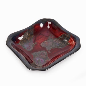 May include: A square ceramic dish with a red glaze and a dark blue rim. The dish has a unique, textured surface with a mottled pattern.