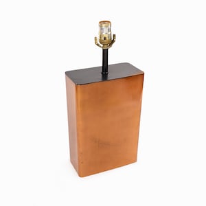 Puede incluir: Una lámpara de mesa de cobre con una base negra y un diseño sencillo. La lámpara tiene una sola bombilla y un cable negro.