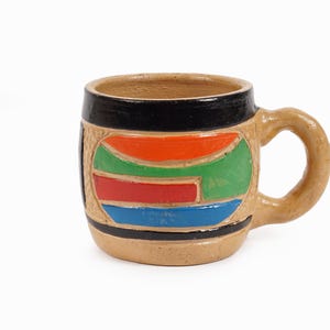 Pode incluir: Uma pequena caneca de cerâmica bege com uma alça. A caneca apresenta uma borda e base pretas, com um design colorido de formas laranja, verdes, vermelhas e azuis. A caneca é provavelmente feita à mão e tem uma aparência rústica.