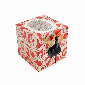 Puede incluir: Una caja decorativa cuadrada con un patrón de remolino rojo y crema. La parte superior presenta un diseño floral. Un cierre negro en forma de libélula con alas ámbar asegura la caja. La caja está sobre un fondo blanco.