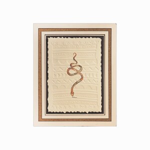 Puede incluir: Una obra de arte enmarcada que presenta una serpiente estilizada con un patrón marrón y naranja sobre un fondo crema con un patrón texturizado. El marco es marrón con un passe-partout crema.