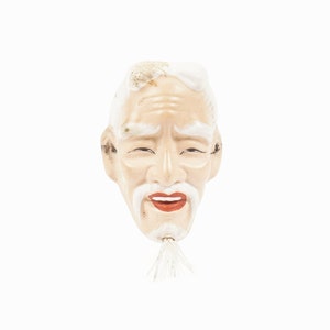 May include: A white ceramic mask of an elderly person with a white beard and mustache. The mask has a painted expression of happiness with a wide smile and closed eyes.