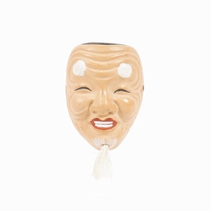 Pode incluir: Uma máscara Noh japonesa tradicional com uma expressão sorridente. A máscara é feita de madeira e pintada em tons de marrom e branco. Tem uma barba branca e círculos brancos ao redor dos olhos.