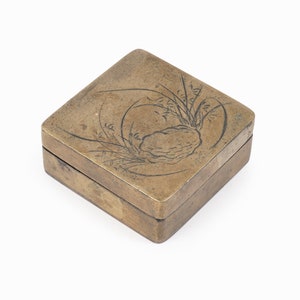 Może przedstawiać: Mała, kwadratowa, mosiężna skrzynka z pokrywką na zawiasach. Pokrywka jest ozdobiona grawerowanym wzorem przedstawiającym skałę i trawę.