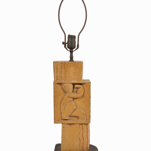 May include: A vintage table lamp with a stacked, geometric design. The lamp features a tan-colored, carved central column with a relief of a seated figure. The base is black, and the lamp has a brown metal shade holder.
