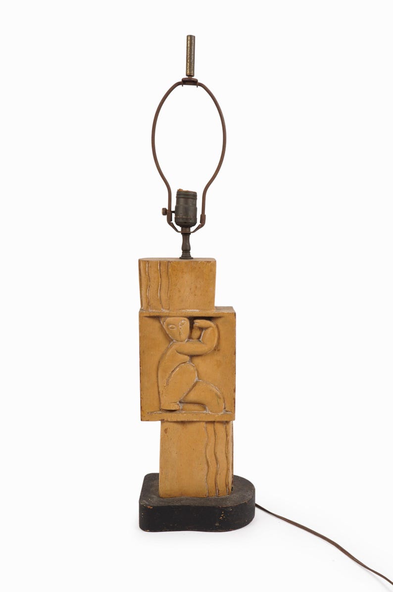 May include: A vintage table lamp with a stacked, geometric design. The lamp features a tan-colored, carved central column with a relief of a seated figure. The base is black, and the lamp has a brown metal shade holder.