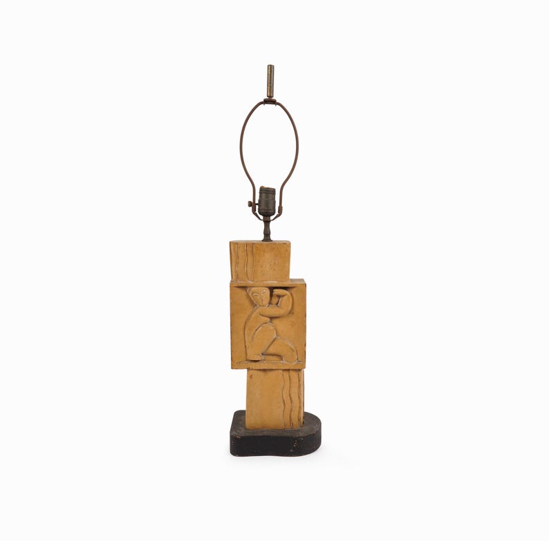 May include: A tan-colored table lamp with a carved relief of a person. The lamp has a rectangular base and a stacked design. The lamp's metal hardware includes a bulb socket and a lamp shade holder.