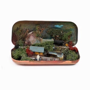 May include: A miniature diorama housed inside an open rectangular metal tin. The scene features a small yellow cabin with a blue roof, green foliage, red shrubs, a tiny white figure, and a black bear figurine set against a painted backdrop.