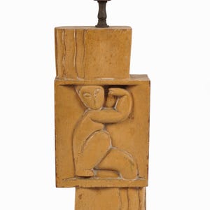 May include: A tan-colored lamp base with a carved relief of a seated figure. The lamp base is rectangular with a carved design and a metal lamp fixture at the top. The figure is in a seated position with one arm raised.