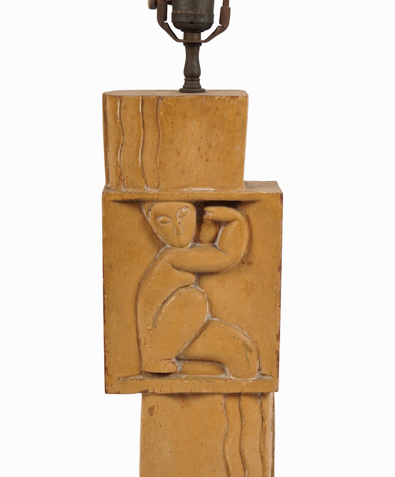 May include: A tan-colored lamp base with a carved relief of a seated figure. The lamp base is rectangular with a carved design and a metal lamp fixture at the top. The figure is in a seated position with one arm raised.