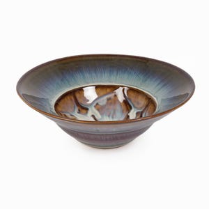 May include: A wide, shallow ceramic bowl featuring a glossy glaze with a deep brown center that transitions into a vibrant blue and dark brown rim. The interior surface displays a marbled, fluid pattern with streaks of white and light blue tones.