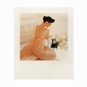 Puede incluir: Una mujer con un vestido de color melocotón con la espalda descubierta se sienta en una silla de mimbre blanca. Está mirando un ramo de tulipanes blancos y melocotón.
