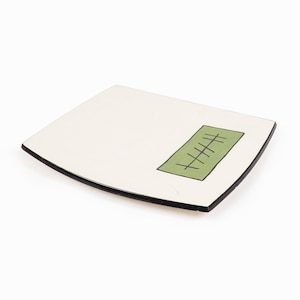 May include: A white ceramic square plate with a black rim and a green rectangle in the center. The rectangle has a series of black lines drawn inside.