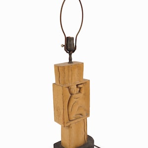 May include: A vintage table lamp with a carved wooden base. The lamp features a rectangular design with a carved figure. The lamp has a brown cord and a metal lamp holder. The base is painted black.