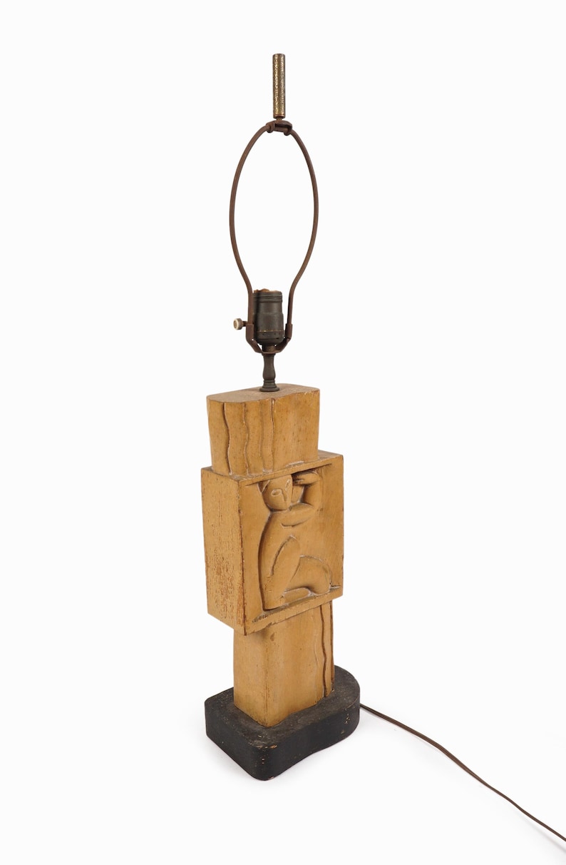 May include: A vintage table lamp with a carved wooden base. The lamp features a rectangular design with a carved figure. The lamp has a brown cord and a metal lamp holder. The base is painted black.