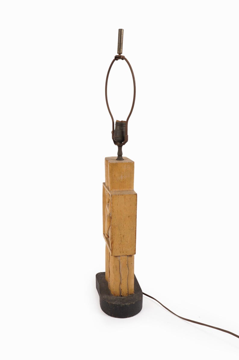 May include: A vintage wooden lamp with a light brown finish. The lamp has a rectangular base and a carved design on the body. The lamp has a metal lamp holder and a brown electrical cord.