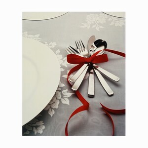 Puede incluir: Un juego de cubiertos atado con un lazo de cinta roja sobre un mantel blanco con estampado floral.