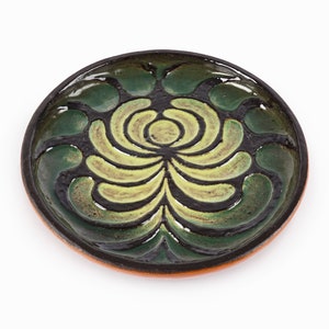 May include: A round ceramic plate with a green and yellow floral design. The plate has a dark brown rim and a light brown base.