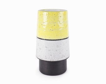 Op de afbeelding: Een gele, witte en zwarte keramische vaas met een textuur. De vaas heeft een brede opening en een smalle basis.