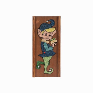May include: A wooden framed wall hanging with a colorful elf holding a shamrock. The elf is made of small colorful beads and is wearing a blue hat, green pants, and blue shoes.