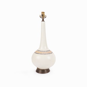 Puede incluir: Lámpara de mesa de cerámica blanca con base bulbosa y cuello esbelto. La lámpara presenta una banda decorativa de rayas doradas, plateadas y naranjas. Tiene un portalámparas color latón y una base de color bronce oscuro.
