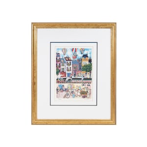 May include: A framed print of a colorful illustration depicting a bustling city street scene with hot air balloons, a train, and people walking and riding bicycles. The print is set against a white background and has a gold frame.