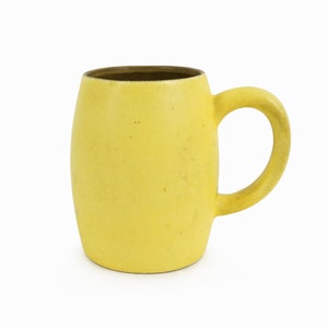 Puede incluir: Una taza de cerámica amarilla con cuerpo redondeado y asa. La taza tiene una textura moteada.