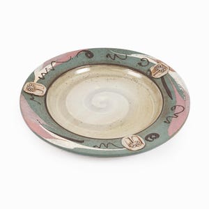 Puede incluir: Un plato de cerámica verde con un diseño de remolino blanco en el centro. El plato tiene un borde rosa y blanco con un diseño marrón y tres diseños de palmeras marrones.