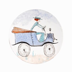 以下が含まれることがあります： 手描きで描かれた、女性がパンを積んだビンテージカーを運転しているイラストが描かれた青と白のセラミックプレートです。
